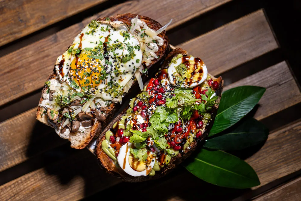 Two open sourdough toasts, one with a fried egg and mushrooms, one with avocado, egg and pomegranate